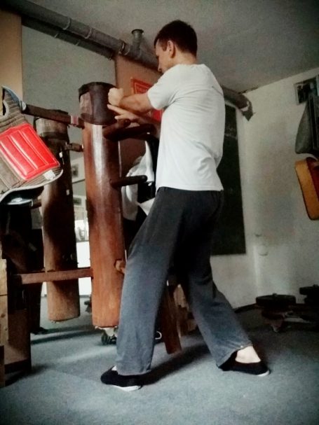 Michael an der Holzpuppe Person in grauem Oberteil und schwarzen Hosen übt an einem Holztrainingstool.
